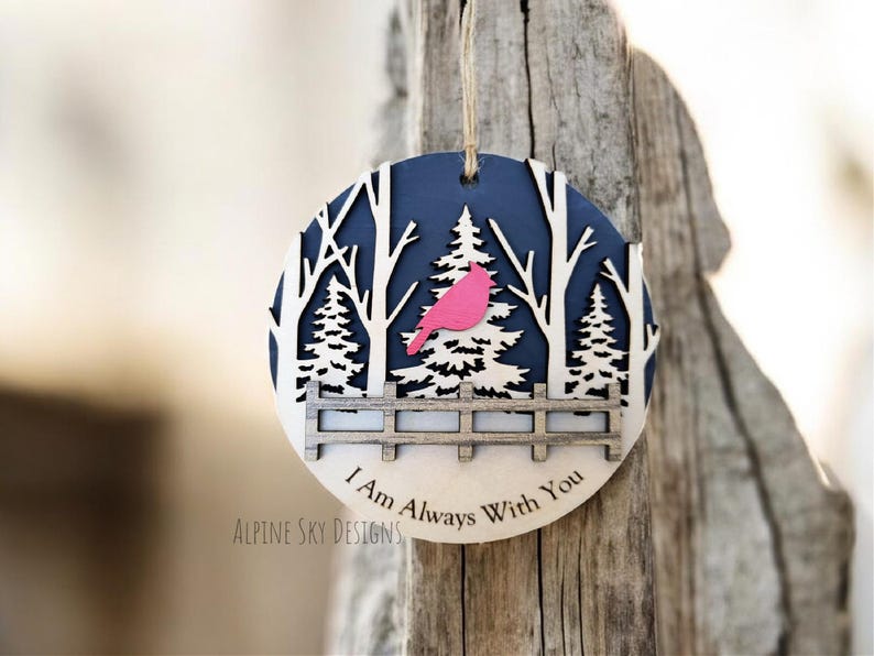 Puede incluir: Un adorno navide&ntilde;o de madera con fondo azul y una silueta blanca de una escena de bosque. Un p&aacute;jaro cardenal rosa est&aacute; posado en una rama. El adorno dice "I Am Always With You" y "Alpine Sky Designs".