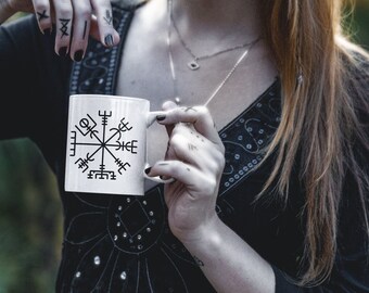 May include: White ceramic mug featuring a black Viking compass symbol. The mug is held by a person with tattoos on their fingers and a dark-colored top. The person has long red hair and is wearing jewelry.