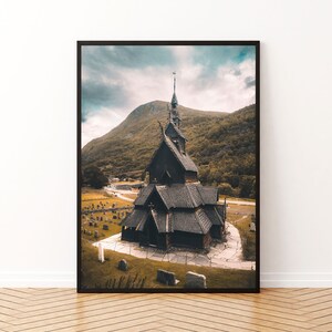 Puede incluir: Impresión enmarcada de una histórica iglesia de madera con una alta aguja, sobre un fondo de montaña y cielo nublado. La iglesia tiene un aspecto oscuro y desgastado, rodeada por un cementerio.