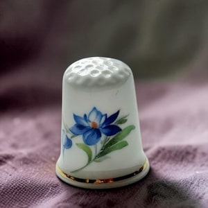May include: A white porcelain thimble featuring a hand-painted blue flower with green leaves on the side. The top of the thimble has a textured dimpled surface, and the base is finished with a thin, reflective gold metallic band trim.
