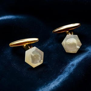 Puede incluir: Un par de gemelos dorados con incrustaciones hexagonales de nácar blanco iridiscente. Cada gemelo tiene una barra de cierre de metal dorado pulido unida a la parte posterior de la cara hexagonal mediante una pequeña bisagra articulada.