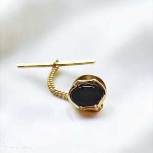 May include: A gold-toned tie tack featuring an oval-shaped dark blue stone set in a metal frame. The piece includes a horizontal bar connected to the main setting by a short, delicate gold-toned chain, designed for securing a necktie in place.