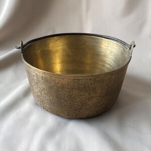 May include: A round brass pot with a weathered, textured exterior and a smooth, polished interior. The vessel features a dark metal swing handle attached to two small, upright lugs on opposite sides of the rim, creating a functional design.