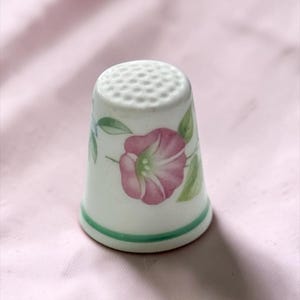 Può includere: Un ditale in ceramica bianca decorato con un fiore di gloria del mattino rosa dipinto a mano, foglie verdi e una sottile fascia verde alla base. La parte superiore del ditale presenta una superficie testurizzata a fossette per il cucito.