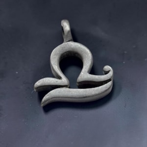 Può includere: Un ciondolo in metallo color argento a forma di simbolo zodiacale della Bilancia. L'oggetto presenta una finitura liscia e opaca con un design curvo e stilizzato e un piccolo occhiello integrato sulla parte superiore per l'aggancio a una catena.