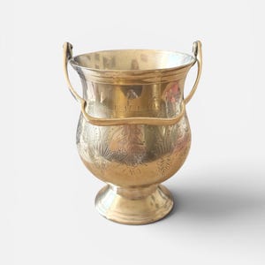 May include: A polished brass bucket with a wide, flared rim and a rounded body. The exterior features intricate floral engravings around the centre. It includes a curved, hinged handle attached to the sides and rests on a circular pedestal base.