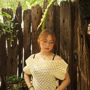 May include: A cream-colored, off-the-shoulder crochet top with short sleeves. The top is paired with a black and white polka dot skirt. The background features a wooden fence and lush greenery.