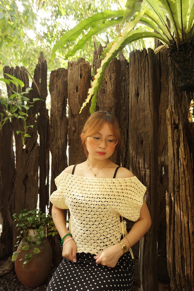 May include: A cream-colored, off-the-shoulder crochet top with short sleeves. The top is paired with a black and white polka dot skirt. The background features a wooden fence and lush greenery.