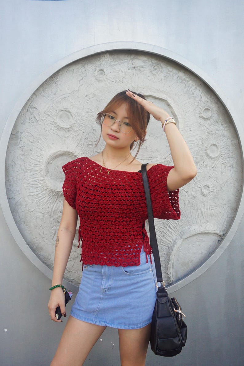 May include: A red crochet top with short sleeves and side ties, paired with a light blue denim skirt. A black shoulder bag is visible. The background features a textured, circular design.