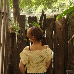 May include: A light yellow, off-the-shoulder crochet top with short sleeves. The top is paired with a black and white polka dot skirt. The person is standing in front of a rustic wooden fence with greenery.