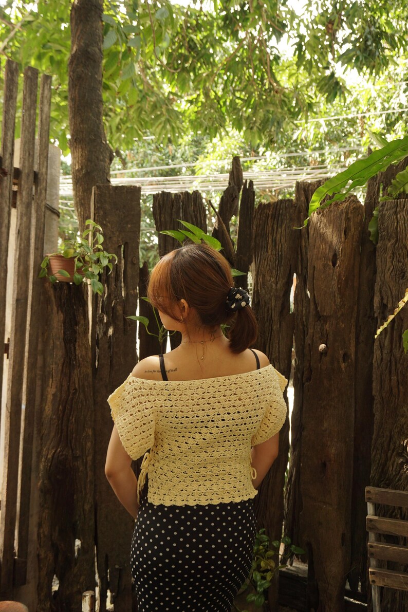 May include: A light yellow, off-the-shoulder crochet top with short sleeves. The top is paired with a black and white polka dot skirt. The person is standing in front of a rustic wooden fence with greenery.