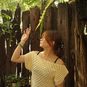 May include: A light yellow, off-the-shoulder crochet top. The top has short sleeves and a textured, open weave design. The person is wearing a black polka dot skirt and a gold watch. The background includes a wooden fence and lush greenery.