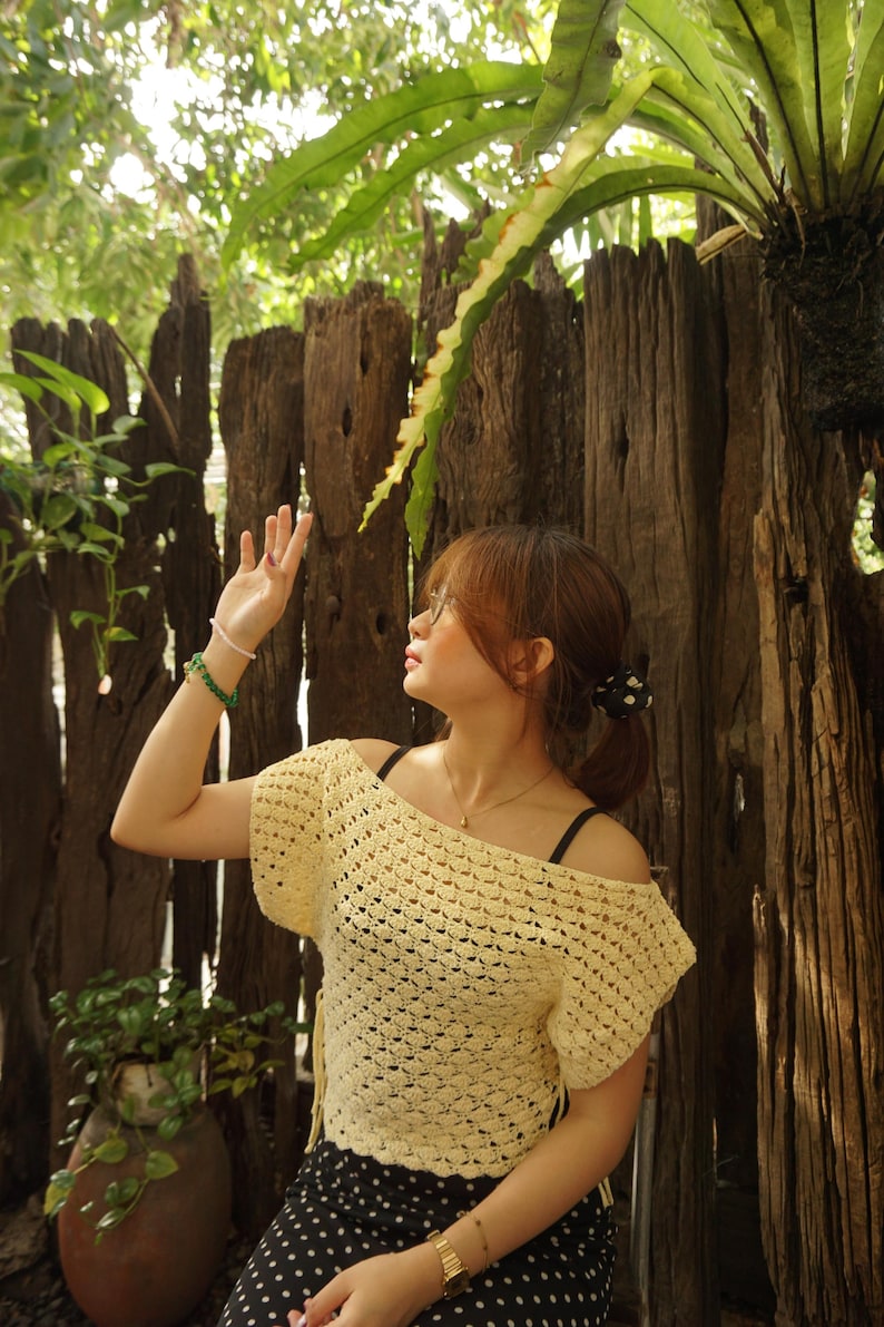 May include: A light yellow, off-the-shoulder crochet top. The top has short sleeves and a textured, open weave design. The person is wearing a black polka dot skirt and a gold watch. The background includes a wooden fence and lush greenery.