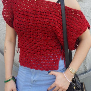 May include: A red, off-the-shoulder crochet top with short sleeves. The top is paired with a denim skirt. A black shoulder bag and gold watch are also visible.