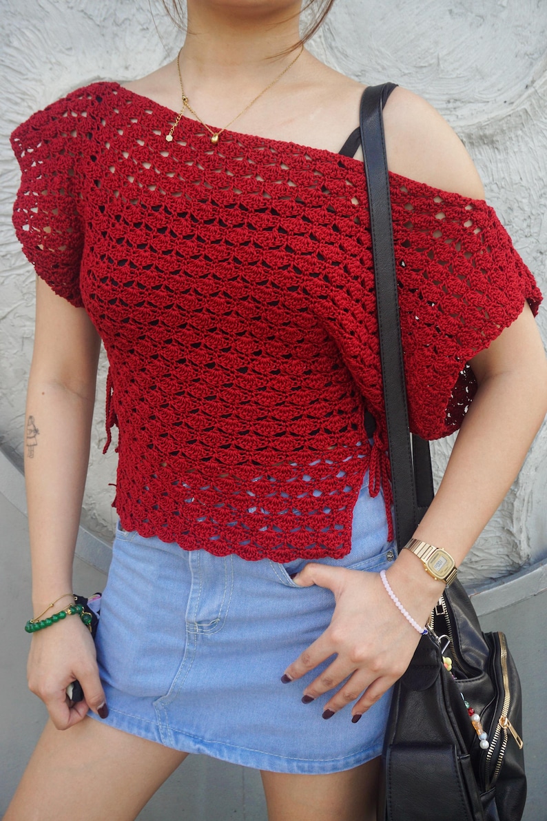 May include: A red, off-the-shoulder crochet top with short sleeves. The top is paired with a denim skirt. A black shoulder bag and gold watch are also visible.