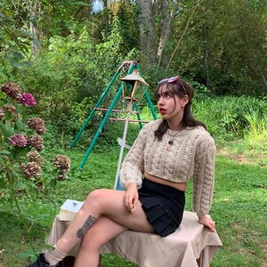 以下が含まれることがあります： ベージュのケーブルニットのクロップドセーター、黒いミニスカート、黒いプラットフォームシューズを履いた女性。彼女は庭のウッドベンチに座っています。