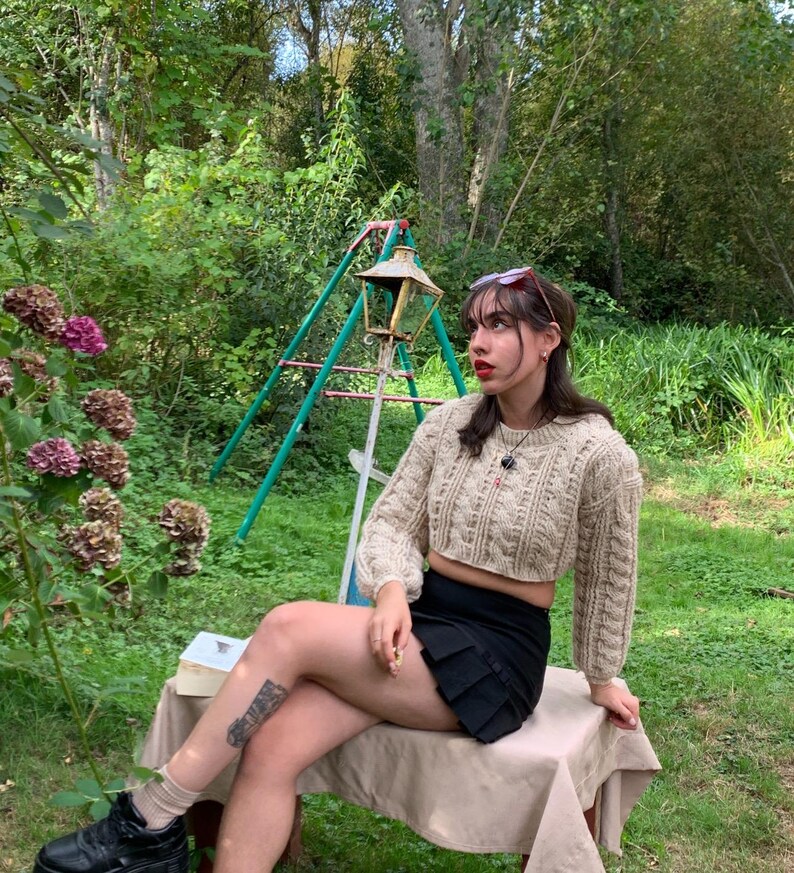 以下が含まれることがあります： ベージュのケーブルニットのクロップドセーター、黒いミニスカート、黒いプラットフォームシューズを履いた女性。彼女は庭のウッドベンチに座っています。