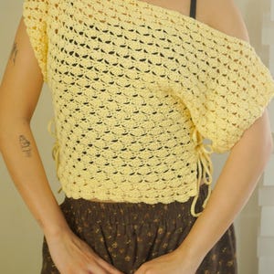 May include: A light yellow, off-the-shoulder crochet top. The top features a scalloped edge and ties on the side. The model is also wearing a brown floral skirt and a gold necklace with charms.