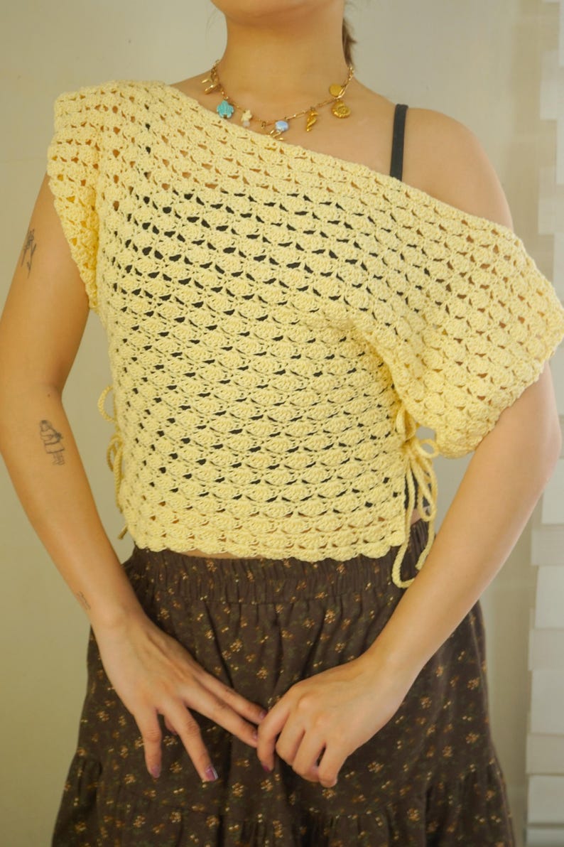 May include: A light yellow, off-the-shoulder crochet top. The top features a scalloped edge and ties on the side. The model is also wearing a brown floral skirt and a gold necklace with charms.