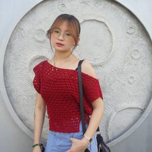 May include: A red crochet top with an off-the-shoulder design. The top is paired with a denim skirt and a black shoulder bag. The background features a textured, circular design.