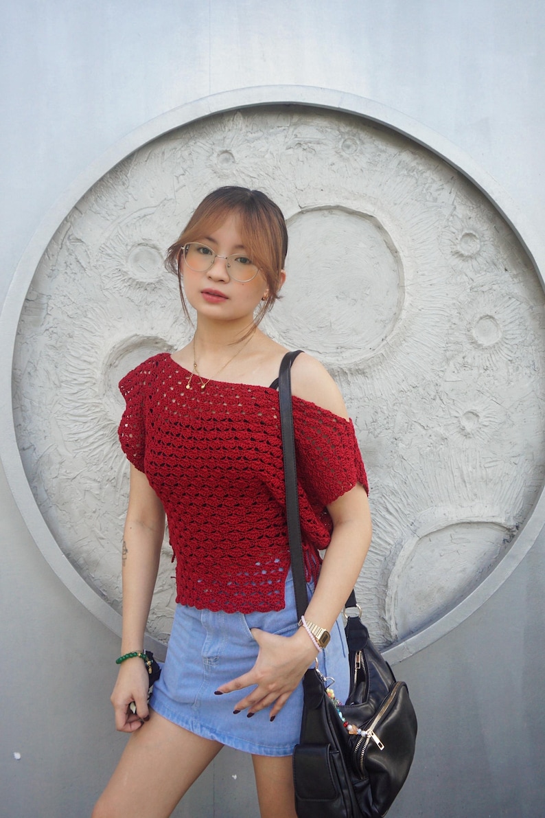 May include: A red crochet top with an off-the-shoulder design. The top is paired with a denim skirt and a black shoulder bag. The background features a textured, circular design.