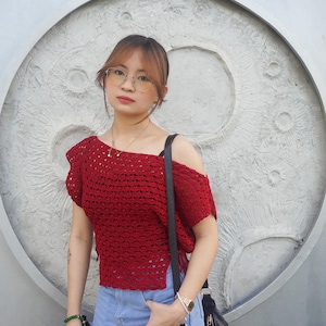 May include: A red crochet top with an off-the-shoulder design. The top is paired with a denim skirt and a black shoulder bag. The background features a textured, circular design.