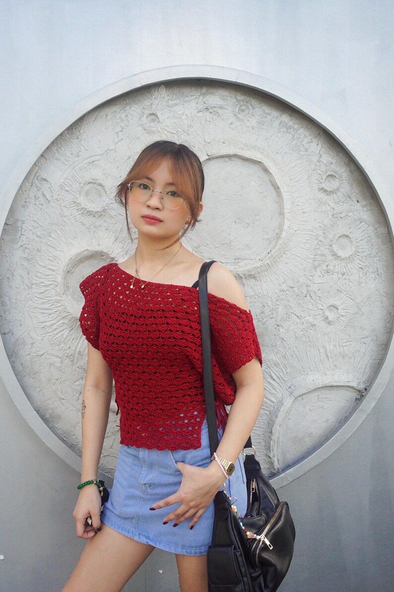 May include: A red crochet top with an off-the-shoulder design. The top is paired with a denim skirt and a black shoulder bag. The background features a textured, circular design.