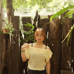 May include: A yellow crochet off-the-shoulder top with short sleeves and tie details. The top is paired with a black and white polka dot skirt. The background includes a wooden fence and lush greenery.