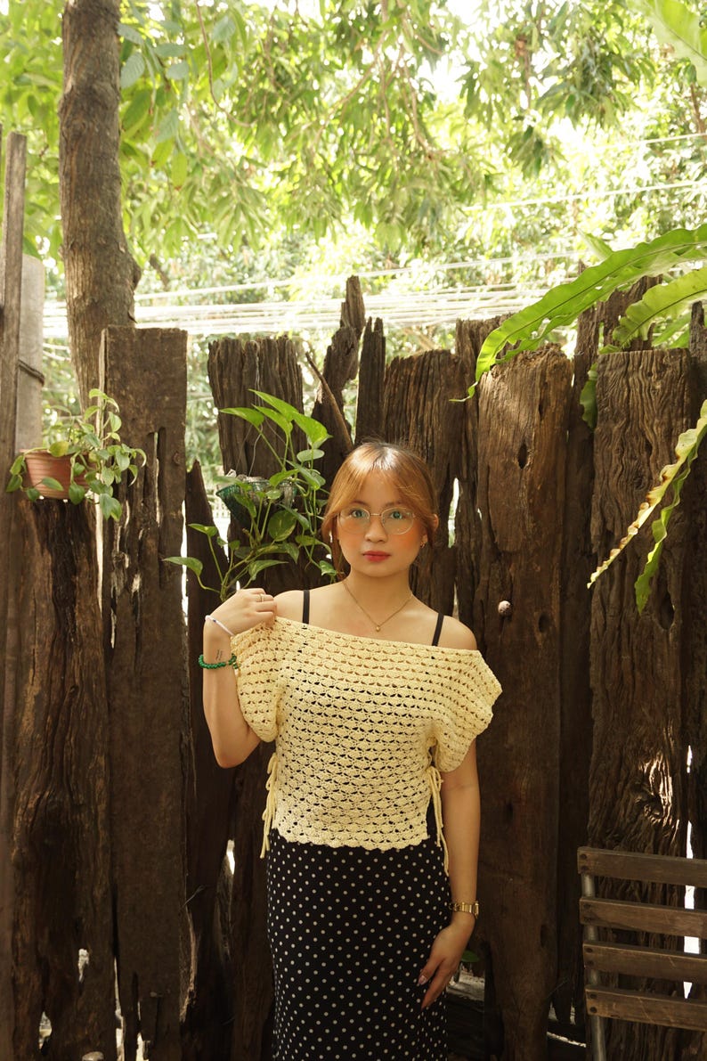 May include: A yellow crochet off-the-shoulder top with short sleeves and tie details. The top is paired with a black and white polka dot skirt. The background includes a wooden fence and lush greenery.