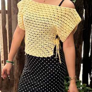 May include: A yellow crochet off-the-shoulder top with short sleeves. The top has a textured, open weave design and is tied at the sides. The top is paired with a black and white polka dot skirt.