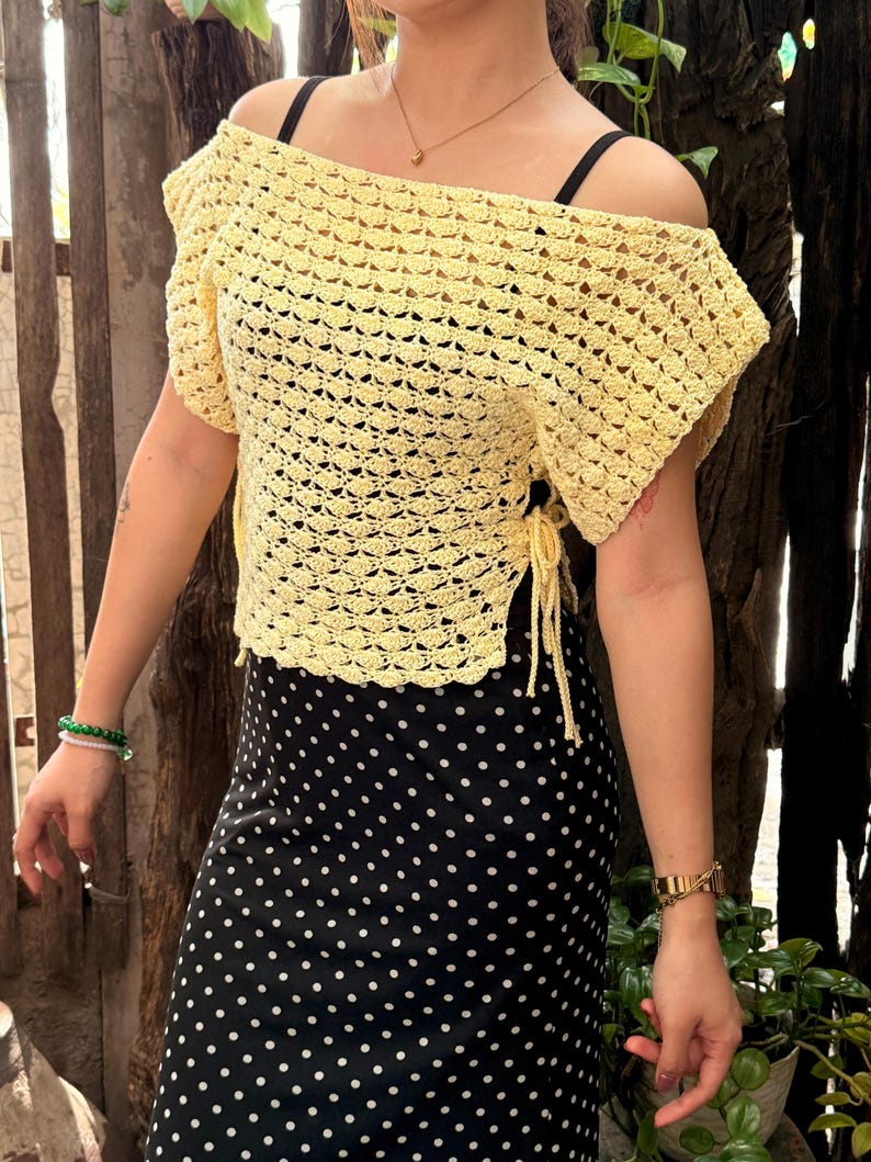 May include: A yellow crochet off-the-shoulder top with short sleeves. The top has a textured, open weave design and is tied at the sides. The top is paired with a black and white polka dot skirt.