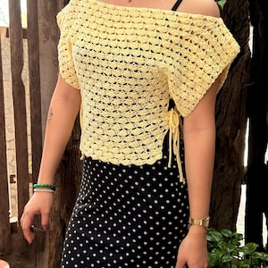 May include: A light yellow, off-the-shoulder crochet top with short sleeves. The top has a textured, open weave design and tie closures on the sides. The model is wearing a black and white polka dot skirt. The top is a handmade item.