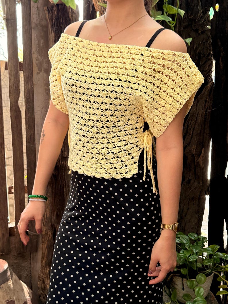 May include: A light yellow, off-the-shoulder crochet top with short sleeves. The top has a textured, open weave design and tie closures on the sides. The model is wearing a black and white polka dot skirt. The top is a handmade item.
