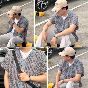 May include: A beige baseball cap with the word "assy" on the front. The person is wearing a short-sleeved, patterned shirt in shades of blue and brown, paired with gray pants. A black bracelet is visible on the wrist.