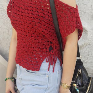 May include: A red crochet top with an off-the-shoulder design and side ties. The top is paired with light blue denim shorts. A black shoulder bag and gold watch are also visible.