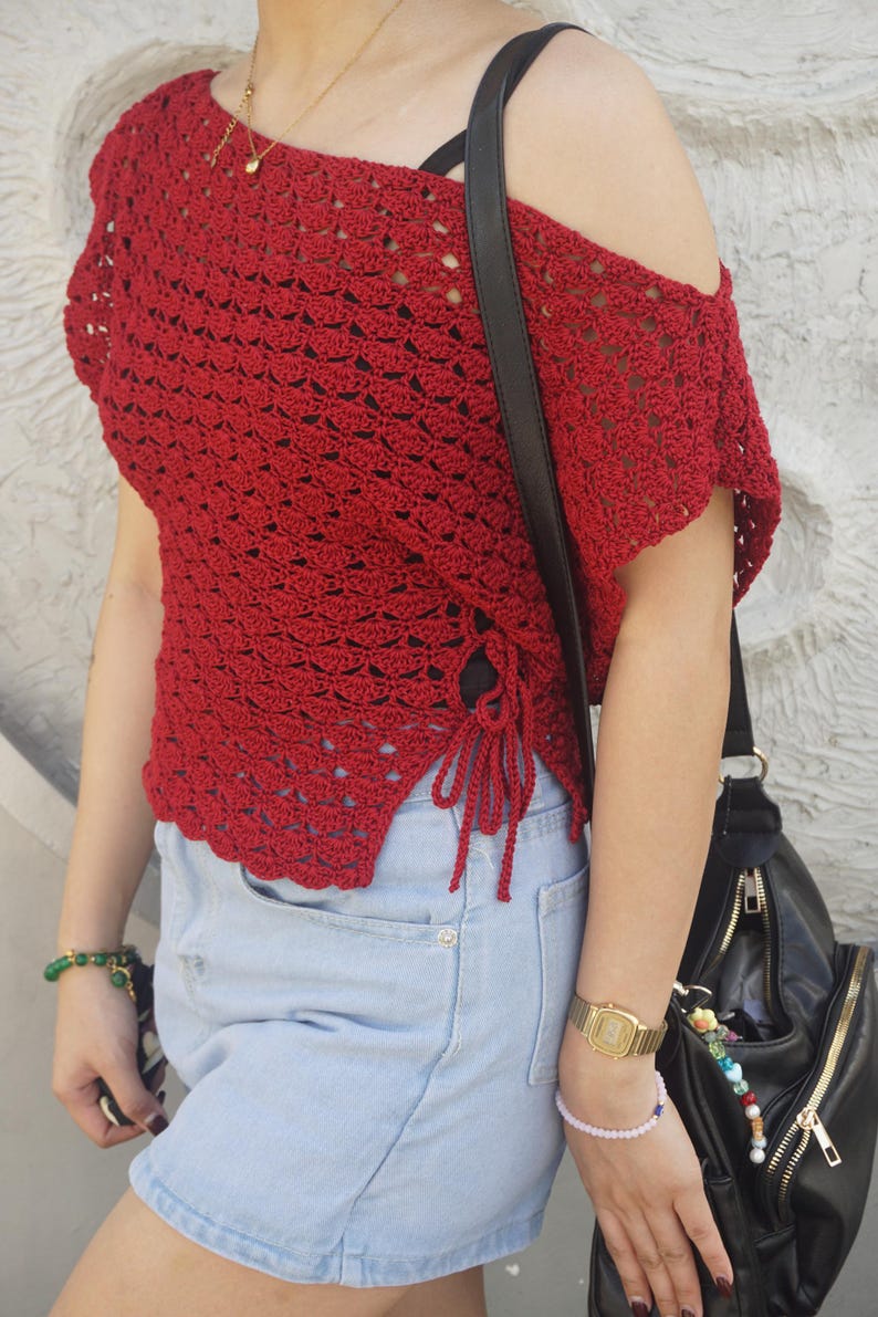 May include: A red crochet top with an off-the-shoulder design and side ties. The top is paired with light blue denim shorts. A black shoulder bag and gold watch are also visible.