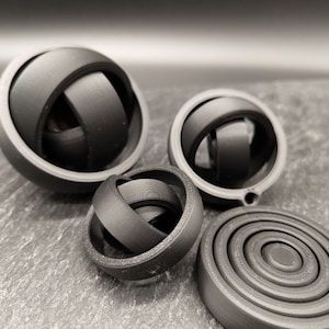 Könnte beinhalten: Vier schwarze, 3D-gedruckte geometrische Formen. Die Formen sind eine Kugel, eine Kugel mit einem Loch, eine Kugel mit einem Loch und einem Ring und eine flache Scheibe mit konzentrischen Kreisen.