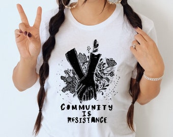 May include: A white t-shirt with a black graphic of two hands holding each other surrounded by flowers. The text "Community is Resistance" is printed below the graphic.