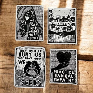 May include: Four black and white stickers with inspirational quotes and illustrations. One features a woman's portrait and the text "I was not made to be subtle." Another says "We rise by lifting others." The third reads "They tried to bury us, they didn't know we were seeds." The last says "Practice radical empathy."