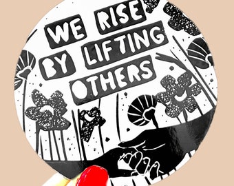 Może przedstawiać: Okrągła naklejka w kolorze czarno-białym z tekstem "We rise by lifting others" w pogrubionych, blokowych literach. Naklejka przedstawia stylizowaną dłoń wyciągniętą w kierunku innej dłoni, otoczoną czarno-białymi wzorami kwiatowymi.