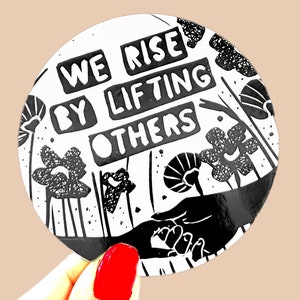 May include: Black and white round sticker with the text "We rise by lifting others" in bold, block letters. The sticker features a stylized hand reaching out to another hand, surrounded by black and white floral designs.