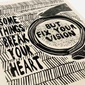 May include: Black and white print with the text "Some things break your heart but fix your vision". The text is in a bold, blocky font and is surrounded by a black and white geometric pattern.