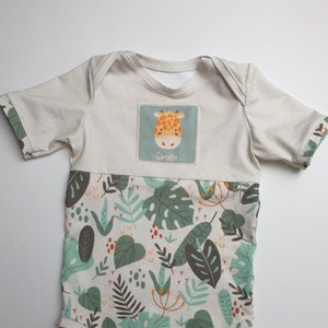 Peut inclure: Une grenouillère bébé de couleur crème à manches courtes avec un imprimé jungle vert, marron et orange. La grenouillère a un patch sur la poitrine avec une girafe de dessin animé et le mot "Giraffe".