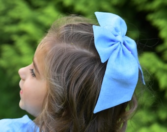 May include: A light blue hair bow is attached to the side of the head of a person with brown curly hair. The person is wearing a light blue top. The background is a blurred green.