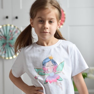 Peut inclure: Un t-shirt blanc avec un motif de fée coloré et une bordure en franges nouées blanches. La fée porte une robe rose et une couronne. La fille porte un pantalon rose.