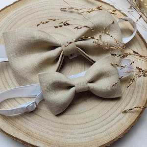 Puede incluir: Dos pajaritas de lino beige con bandas el&aacute;sticas blancas. Las pajaritas est&aacute;n sobre una superficie de madera con hierba seca.