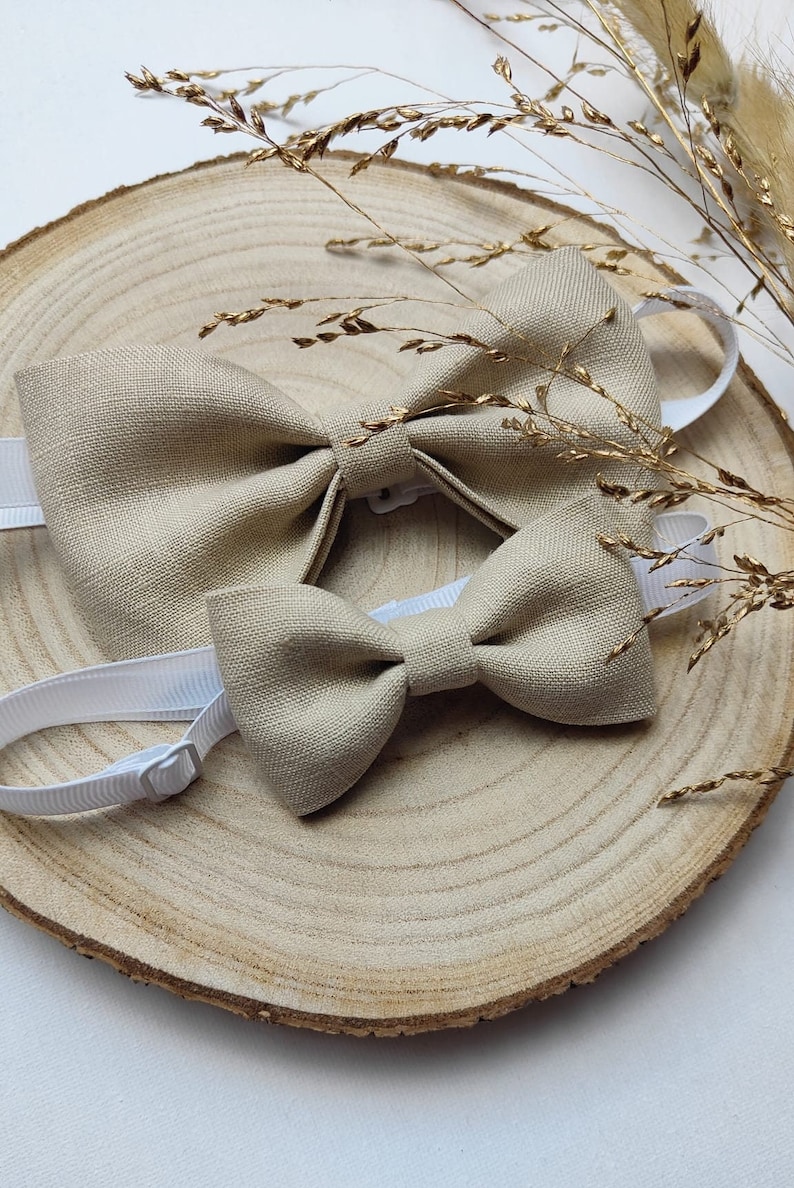 Puede incluir: Dos pajaritas de lino beige con bandas el&aacute;sticas blancas. Las pajaritas est&aacute;n sobre una superficie de madera con hierba seca.