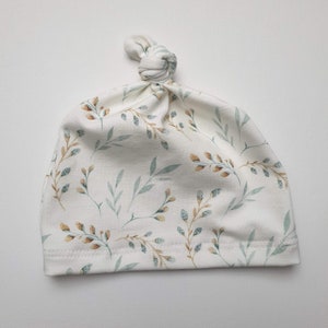Peut inclure: Un bonnet blanc pour b&eacute;b&eacute; avec un n&oelig;ud sur le dessus. Le bonnet est orn&eacute; d'un motif de petites feuilles vertes et brunes.