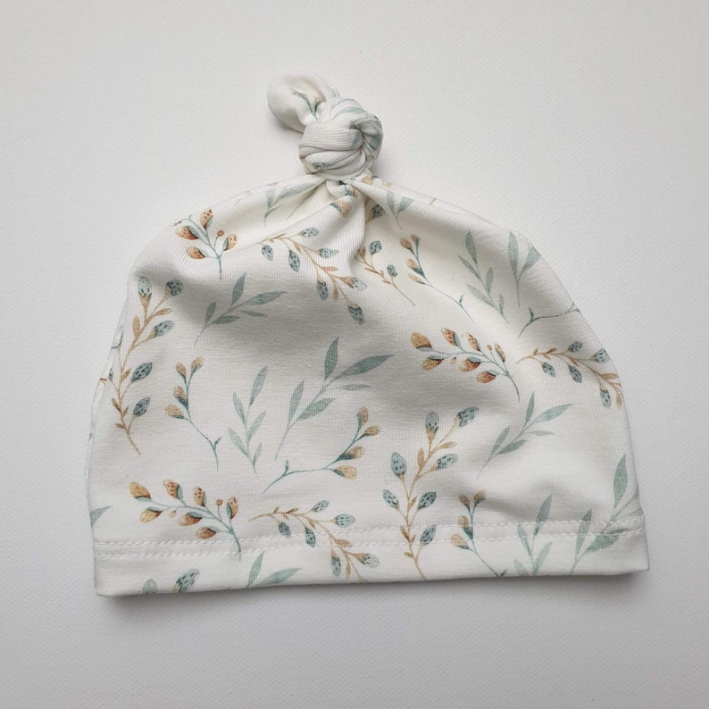 Peut inclure: Un bonnet blanc pour b&eacute;b&eacute; avec un n&oelig;ud sur le dessus. Le bonnet est orn&eacute; d'un motif de petites feuilles vertes et brunes.
