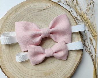 May include: Two pink linen bow ties with white ribbon ties. The bow ties are on a wooden slice.